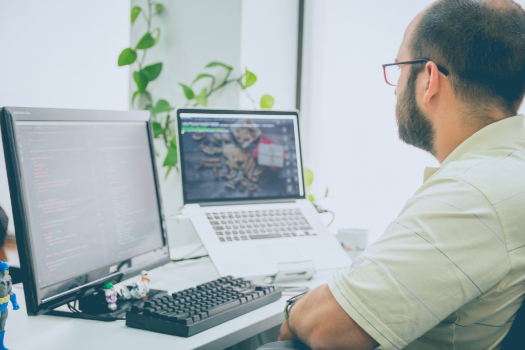 Programmer in a bright office with 2 computer screens looking at one.